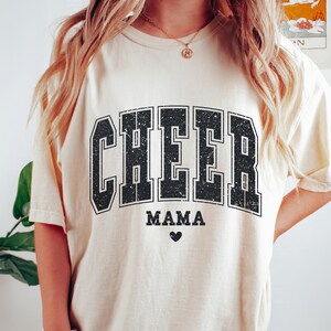 May include: A cream colored t-shirt with a black distressed graphic that says "CHEER MAMA" with a small heart below the text.