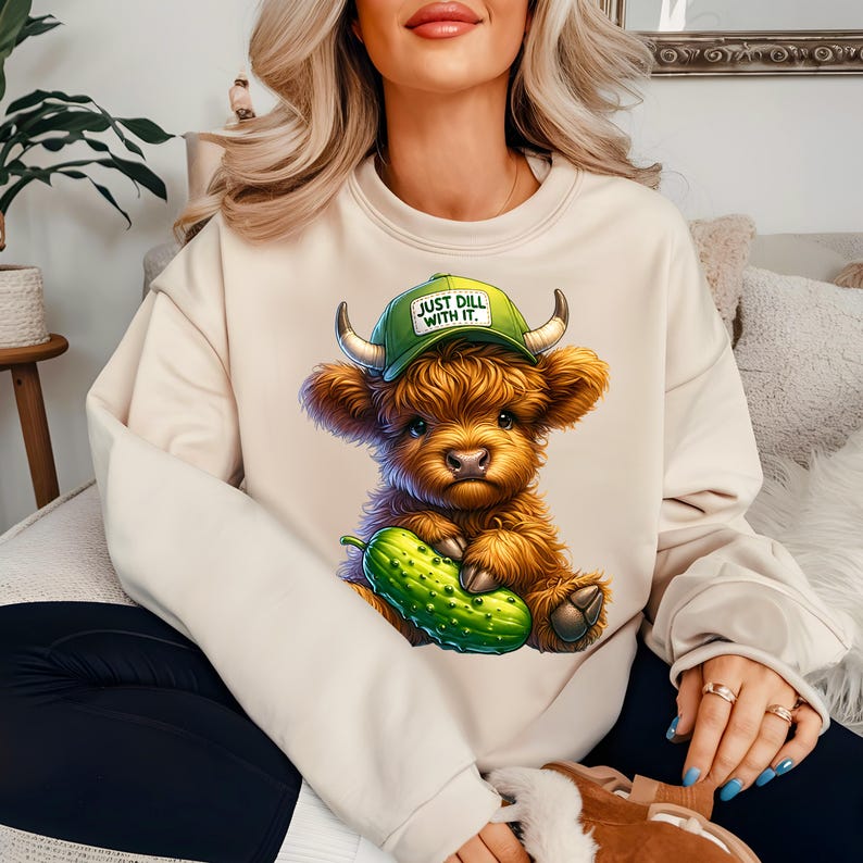 May include: A cream colored sweatshirt with a cartoon highland cow wearing a green and white baseball cap that says "Just Dill With It." The cow is holding a green pickle.