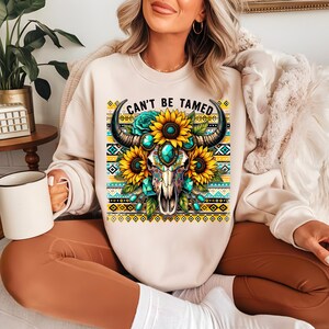 K&ouml;nnte beinhalten: Ein cremefarbener Pullover mit einem Motiv eines Sch&auml;dels mit H&ouml;rnern, umgeben von Sonnenblumen und t&uuml;rkisfarbenen Juwelen. Der Text "Can't Be Tamed" ist &uuml;ber dem Sch&auml;del gedruckt.
