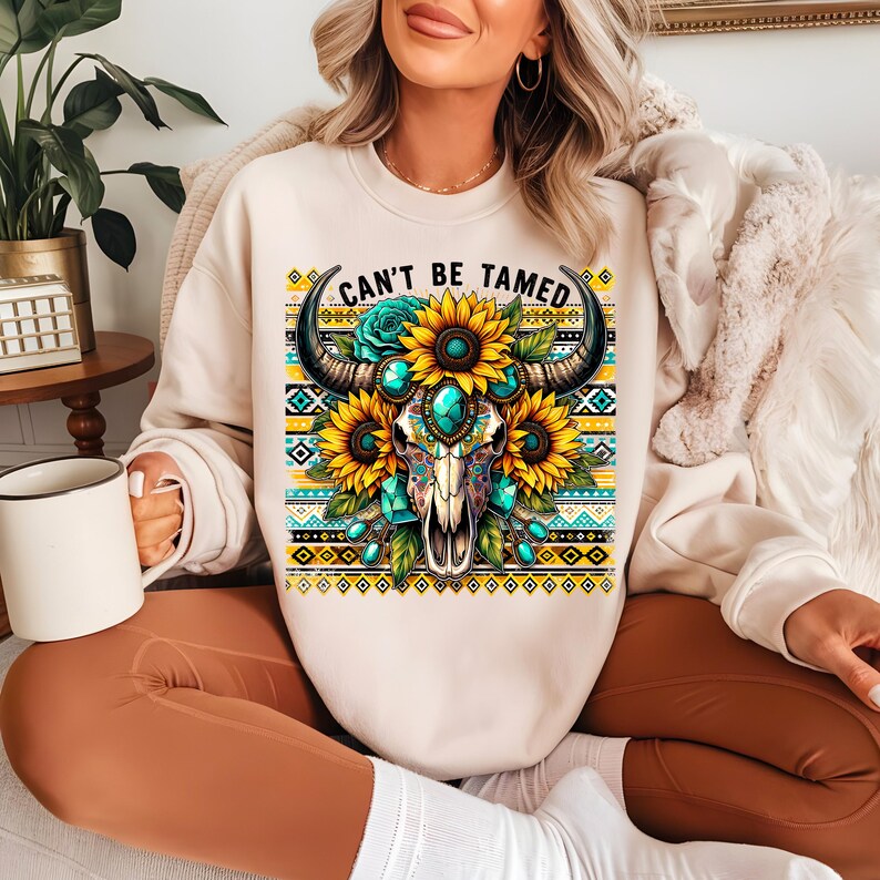 K&ouml;nnte beinhalten: Ein cremefarbener Pullover mit einem Motiv eines Sch&auml;dels mit H&ouml;rnern, umgeben von Sonnenblumen und t&uuml;rkisfarbenen Juwelen. Der Text "Can't Be Tamed" ist &uuml;ber dem Sch&auml;del gedruckt.