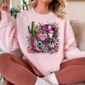 May include: Pink sweatshirt with a graphic design of a cactus, a cowboy hat, a skull, a boot, and flowers. The text "Just a small town girl" is on a pink heart.
