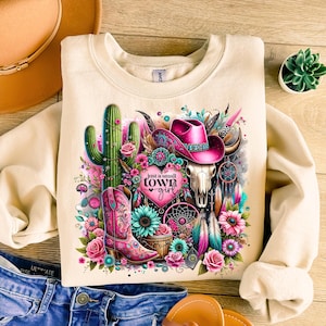 May include: A beige sweatshirt with a colorful graphic design featuring a cactus, a skull, a cowboy hat, feathers, and a boot. The graphic also includes the text "Just a small town girl" with a heart.