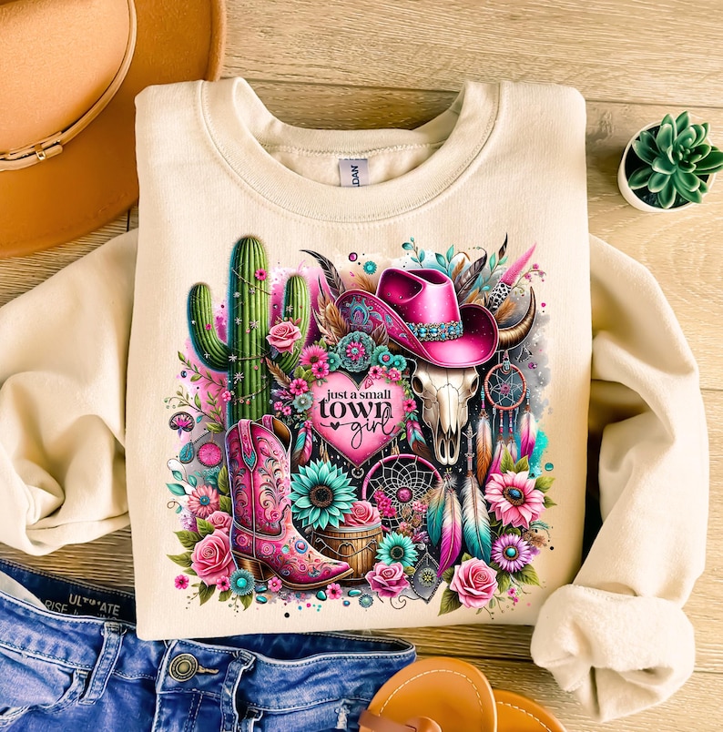 May include: A beige sweatshirt with a colorful graphic design featuring a cactus, a skull, a cowboy hat, feathers, and a boot. The graphic also includes the text "Just a small town girl" with a heart.