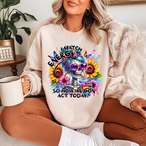 May include: A white sweatshirt with a colorful graphic of a skeleton wearing sunglasses and a headband, holding a drink. The text on the sweatshirt reads "I Match Energy So How We Gon' Act Today?"