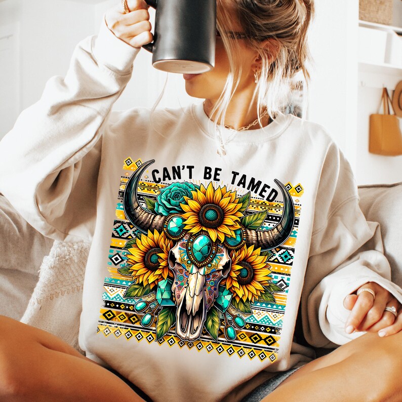 K&ouml;nnte beinhalten: Wei&szlig;er Pullover mit einem bunten Totenkopf-Design und dem Text "Can't Be Tamed". Der Totenkopf ist von Sonnenblumen und t&uuml;rkisfarbenen Edelsteinen umgeben. Der Hintergrund zeigt ein geometrisches Muster in Gelb, Blau und Wei&szlig;.