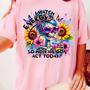 May include: Pink t-shirt with a graphic of a skeleton wearing sunglasses and a bandana, holding a drink. The text reads "I Match Energy So How We Gon' Act Today?"
