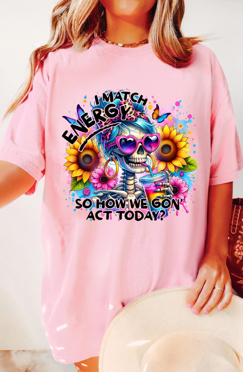 May include: Pink t-shirt with a graphic of a skeleton wearing sunglasses and a bandana, holding a drink. The text reads "I Match Energy So How We Gon' Act Today?"