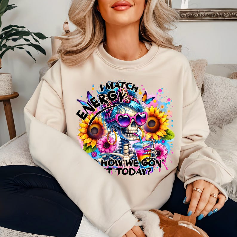 May include: A beige sweatshirt with a colorful graphic of a skeleton wearing sunglasses and holding a drink. The graphic includes the text "I Match Energy How We Gon' Do It Today?"