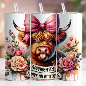 Pode incluir: Um copo branco com um laço rosa e uma vaca Highland com o texto "Aparentemente eu tenho uma atitude". O copo é rodeado por flores rosa e brancas.