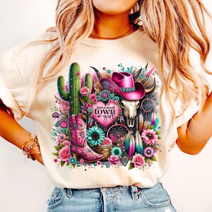 May include: A white t-shirt with a pink and green graphic design featuring a cactus, a cowboy hat, a skull, and a dreamcatcher. The text "just a small town girl" is printed on the shirt.
