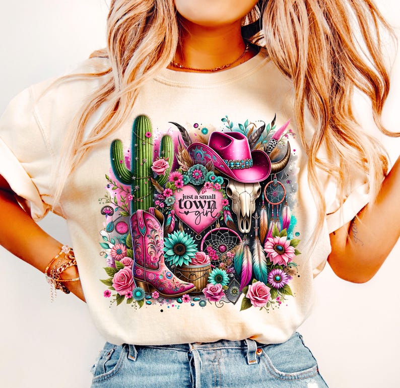 May include: A white t-shirt with a pink and green graphic design featuring a cactus, a cowboy hat, a skull, and a dreamcatcher. The text "just a small town girl" is printed on the shirt.