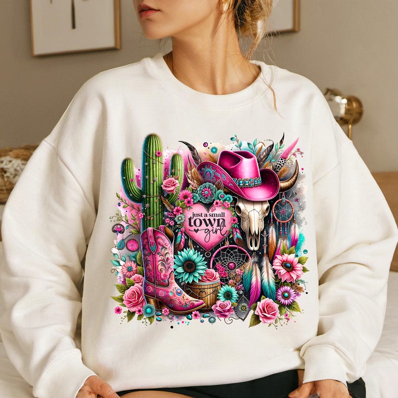 May include: White sweatshirt with a colorful graphic print of a cactus, cowboy hat, boots, feathers, and a skull. The text "Just a small town girl" is printed on the graphic.
