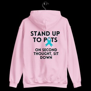 Pode incluir: Moletom rosa claro com o texto "STAND UP TO POTS" acima de uma fita azul-turquesa e a frase "ON SECOND THOUGHT, SIT DOWN" abaixo. O moletom est&aacute; pendurado em um cabide preto contra um fundo preto.