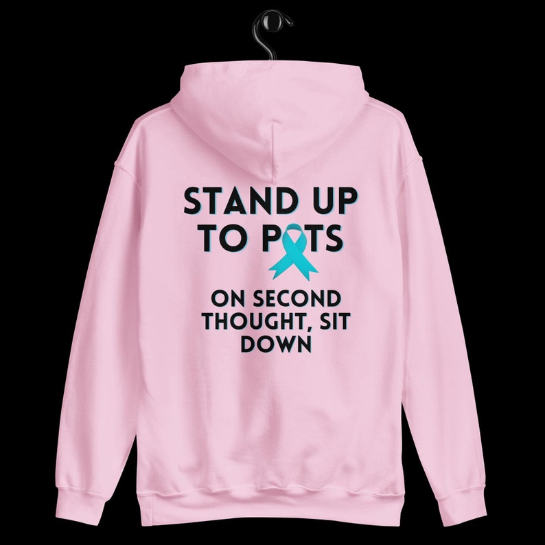 Pode incluir: Moletom rosa claro com o texto "STAND UP TO POTS" acima de uma fita azul-turquesa e a frase "ON SECOND THOUGHT, SIT DOWN" abaixo. O moletom est&aacute; pendurado em um cabide preto contra um fundo preto.