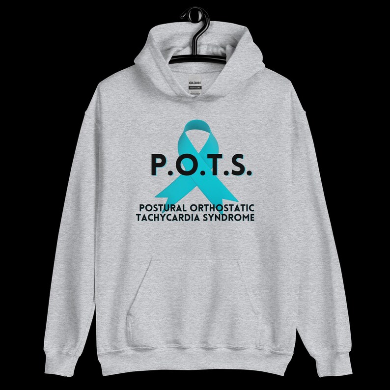 Pode incluir: Um moletom cinza claro com um bolso frontal e um capuz. A frente do moletom apresenta o texto "P.O.T.S." acima de uma fita azul-turquesa, com as palavras "POSTURAL ORTHOSTATIC TACHYCARDIA SYNDROME" abaixo.