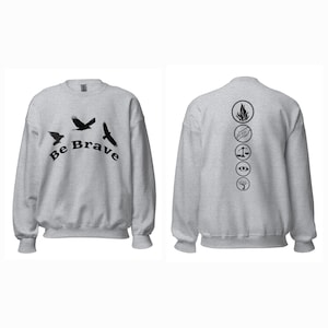 Puede incluir: Una sudadera gris claro con gráficos negros. La parte delantera presenta tres pájaros volando y las palabras "Be Brave". La parte trasera muestra símbolos circulares. Una parte superior cómoda e informal.