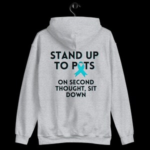 Pode incluir: Um moletom cinza claro com o texto "STAND UP TO POTS" acima de uma fita azul-turquesa e a frase "ON SECOND THOUGHT, SIT DOWN". O moletom est&aacute; pendurado em um cabide preto.