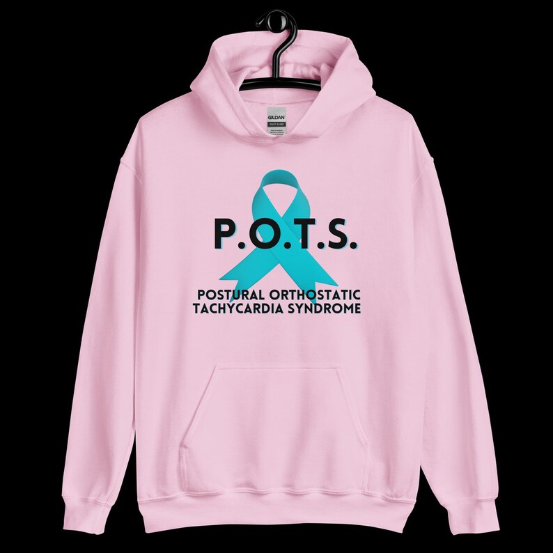 Pode incluir: Moletom rosa claro com um la&ccedil;o turquesa e o texto "P.O.T.S." acima de "Postural Orthostatic Tachycardia Syndrome" em preto. O moletom tem um bolso frontal e um capuz com cord&atilde;o.