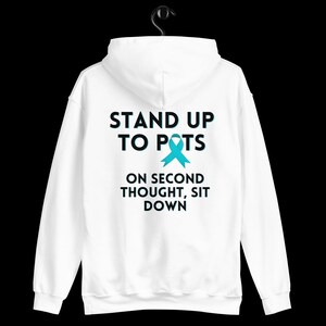 Pode incluir: Moletom branco com a frase "STAND UP TO POTS" acima de uma fita azul-petr&oacute;leo e a frase "ON SECOND THOUGHT, SIT DOWN". O moletom est&aacute; pendurado em um cabide preto em um fundo preto.