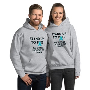 Könnte beinhalten: Zwei hellgraue Kapuzenpullover mit dem Text "STAND UP TO POTS" und "ON SECOND THOUGHT, SIT DOWN" in schwarzer Schrift. Eine türkisfarbene Schleife befindet sich über dem Text. Die Kapuzenpullover haben eine Vordertasche und eine Kapuze.