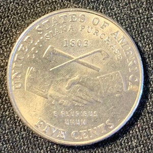 May include: A silver US five-cent coin with the inscription "United States of America" and "Five Cents" around the edge. The center of the coin features a depiction of the 1803 Louisiana Purchase with the text "Louisiana Purchase 1803" and a handshake.