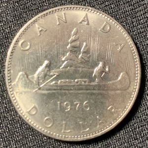 Könnte beinhalten: Eine kanadische Ein-Dollar-Münze aus Silber mit der Darstellung von zwei Personen in einem Kanu, die durch einen Wald paddeln. Das Jahr 1976 ist auf der Münze eingeprägt.