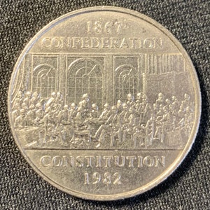 Op de afbeelding: Een zilveren munt ter herdenking van de Confederatie van Canada van 1867 met de tekst "1867 Confederation Constitution 1982" en een afbeelding van mensen die in een kamer bijeengekomen zijn.