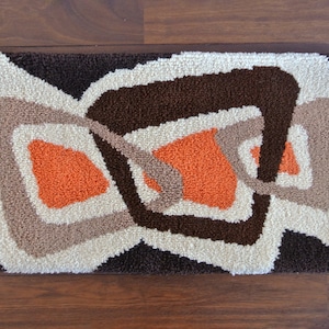 May include: Rectangular rug with an abstract design in shades of brown, beige, and orange. The rug features a repeating geometric pattern with a textured, tufted appearance, suitable for a living room or bedroom.