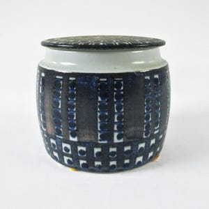 Op de afbeelding: Een witte keramische pot met een deksel, met een geometrisch patroon van zwarte en blauwe cirkels en vierkanten. De pot heeft een ronde vorm en een licht verhoogde basis.