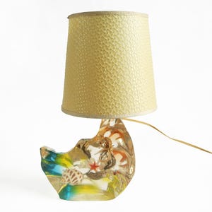 Puede incluir: Lámpara de mesa decorativa con base temática de conchas marinas y una pantalla texturizada de color amarillo claro. La base presenta una forma de media luna con conchas incrustadas, una estrella roja y detalles coloridos. La lámpara tiene un cable dorado.