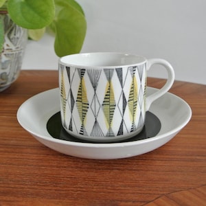 May include: A white ceramic cup and saucer set with a geometric diamond pattern in black, yellow, and gray. The cup sits on a white saucer with a dark green center. A potted plant is in the background.