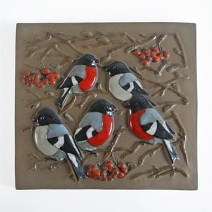 Könnte beinhalten: Quadratische, braune Keramikfliese mit einem Reliefdesign von fünf Vögeln mit roten Brüsten, grauen Flügeln und schwarzen Köpfen. Die Vögel sitzen auf Ästen mit roten Beeren. Der Hintergrund ist ein strukturiertes Braun.