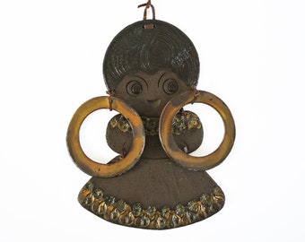 May include: A ceramic wall hanging of a stylized person with large, round, gold earrings. The person has a brown face, black hair, and a brown dress with gold accents.