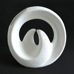 Puede incluir: Un jarrón de cerámica blanca con un marco exterior circular en forma de anillo y dos elementos interiores curvos y puntiagudos que se elevan desde la base. La superficie del objeto presenta un acabado granulado y texturizado muy distintivo.