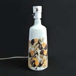 Può includere: Lampada da tavolo in ceramica bianca con un motivo floreale nei toni del marrone e dell'oro. La lampada ha una forma cilindrica e un cavo bianco. La lampada è su sfondo nero.