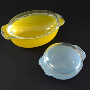 May include: Two vintage Pyrex casserole dishes with clear glass lids. One is a bright yellow color, and the other is a light blue. Both have handles and are on a black surface. These are classic kitchenware items.