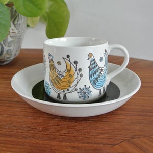 May include: White ceramic mug and saucer set with a bird design in yellow, blue, and black. The mug sits on a white saucer with a dark green center, placed on a wooden surface. A decorative plant is in the background.