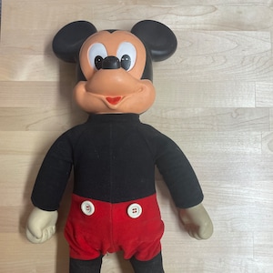 Könnte beinhalten: Ein Vintage-Plüschtier Mickey Mouse mit schwarzem Fell, roten Shorts und gelben Schuhen. Das Spielzeug hat ein klassisches Mickey Mouse-Gesicht mit schwarzen Augen, einer roten Nase und einem breiten Lächeln.