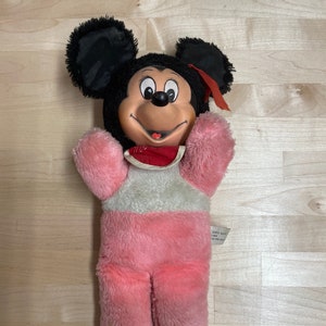 Peut inclure: Un jouet en peluche Mickey Mouse rose et gris avec des oreilles noires et un n&oelig;ud rouge. Le jouet a un visage souriant avec des yeux noirs et une bouche rouge.
