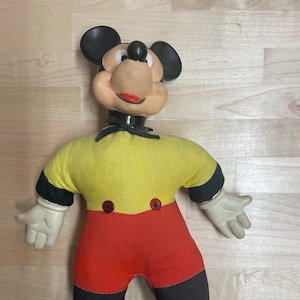 May include: A vintage plush Mickey Mouse doll with black ears, a yellow shirt, red pants, and black shoes. The doll has a black bow tie and a friendly expression.