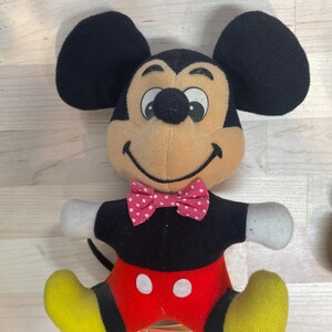 Puede incluir: Un peluche de Mickey Mouse con orejas negras, cara color canela y pajarita de lunares rojos y blancos. El juguete tiene una camisa negra, pantalones cortos rojos con botones blancos, pies amarillos y guantes blancos. Una etiqueta est&aacute; adjunta.