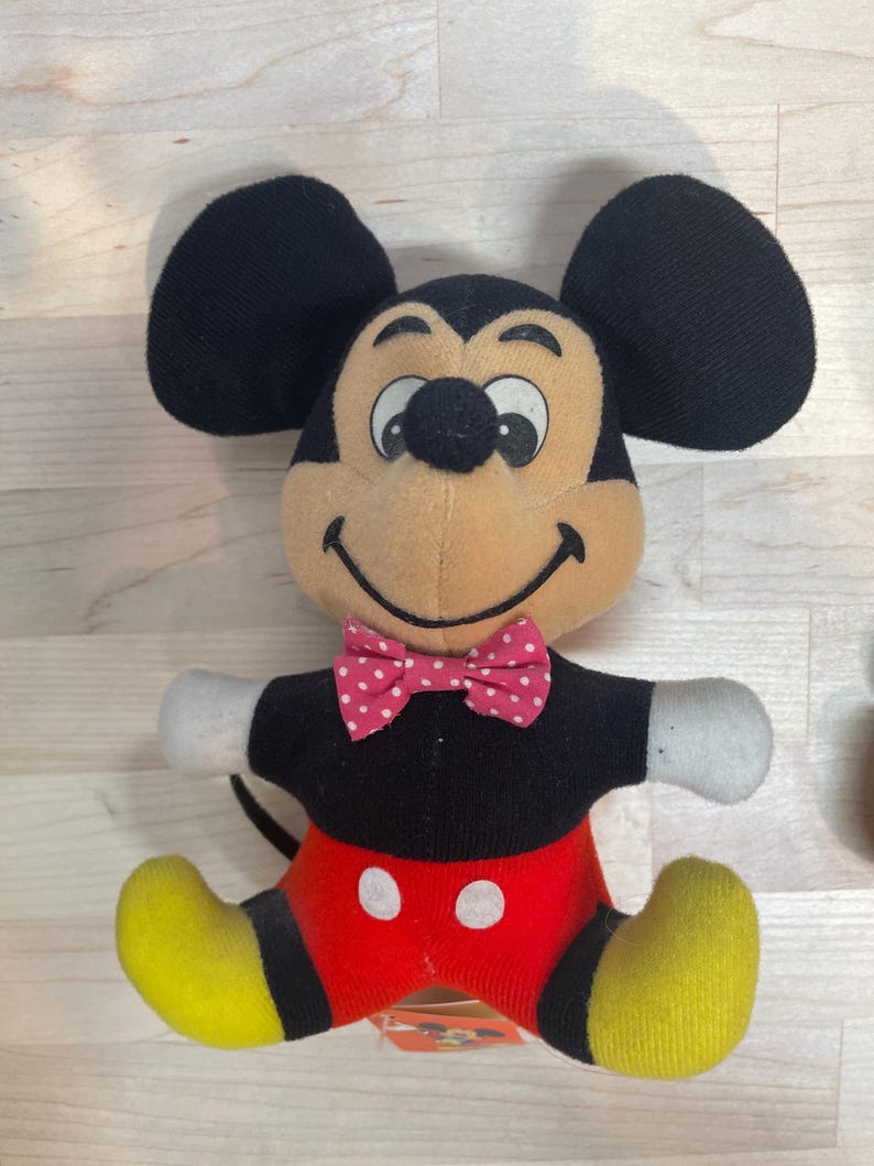 Puede incluir: Un peluche de Mickey Mouse con orejas negras, cara color canela y pajarita de lunares rojos y blancos. El juguete tiene una camisa negra, pantalones cortos rojos con botones blancos, pies amarillos y guantes blancos. Una etiqueta est&aacute; adjunta.