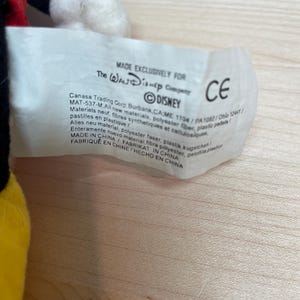 May include: A close-up of a white tag with text, including "MADE EXCLUSIVELY FOR The Walt Disney Company" and the Disney logo. The tag also includes the CE mark and other text in multiple languages. The tag is attached to a plush toy.