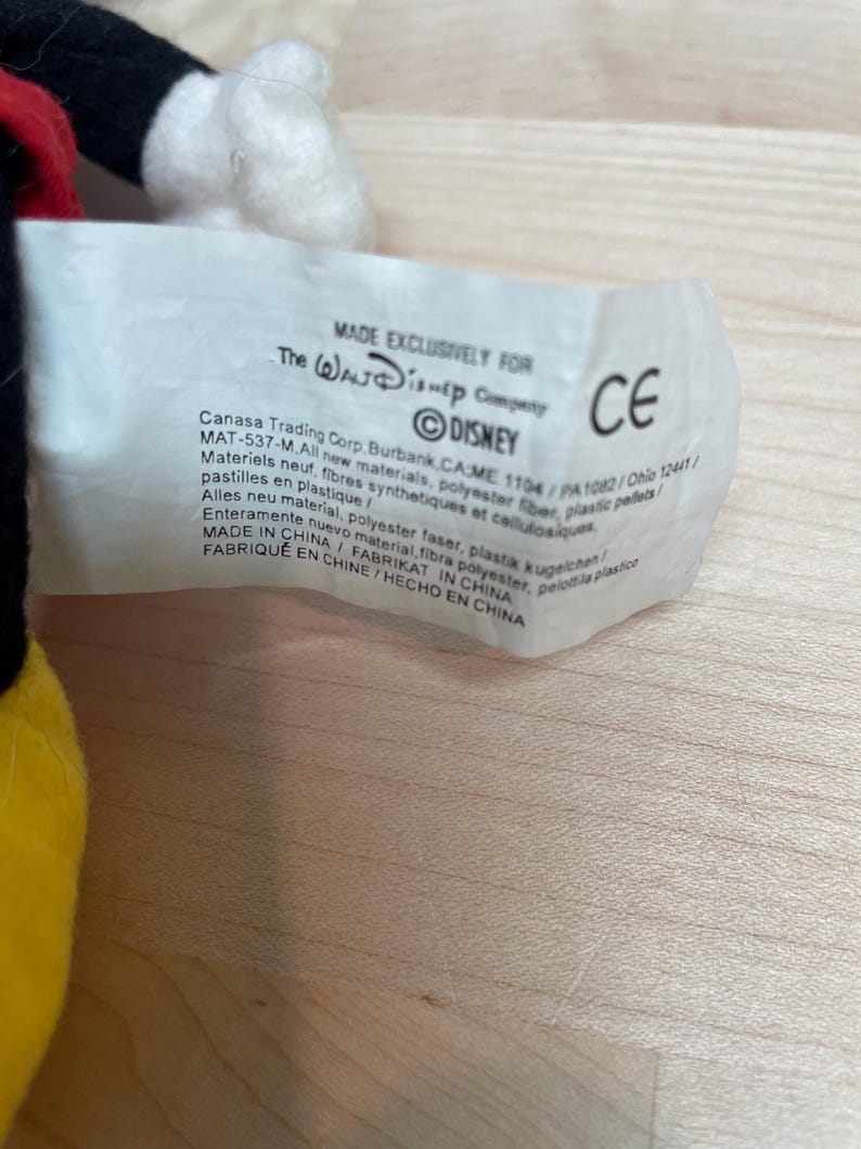 May include: A close-up of a white tag with text, including "MADE EXCLUSIVELY FOR The Walt Disney Company" and the Disney logo. The tag also includes the CE mark and other text in multiple languages. The tag is attached to a plush toy.