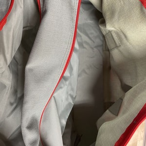 Puede incluir: El interior de una bolsa de mano con cremallera gris y roja. La bolsa est&aacute; hecha de un tejido suave y tejido. La cremallera es roja y recorre la parte superior de la bolsa.