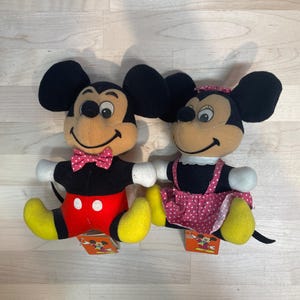 Puede incluir: Dos mu&ntilde;ecos de peluche de Mickey y Minnie Mouse. Mickey lleva pantalones cortos rojos con botones blancos, una camisa negra y una pajarita rosa. Minnie lleva un vestido rosa con lunares, una blusa negra y un lazo rosa. Ambos tienen pies amarillos y orejas negras.