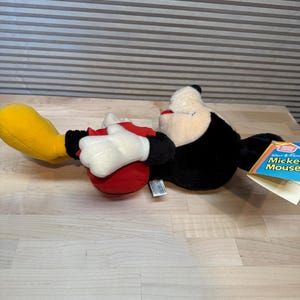 May include: A Mickey Mouse soft toy, lying on its side. The toy has a black body, white gloves, red shorts, and yellow shoes. A tag with the words "Walt Disney Mickey Mouse" is attached.