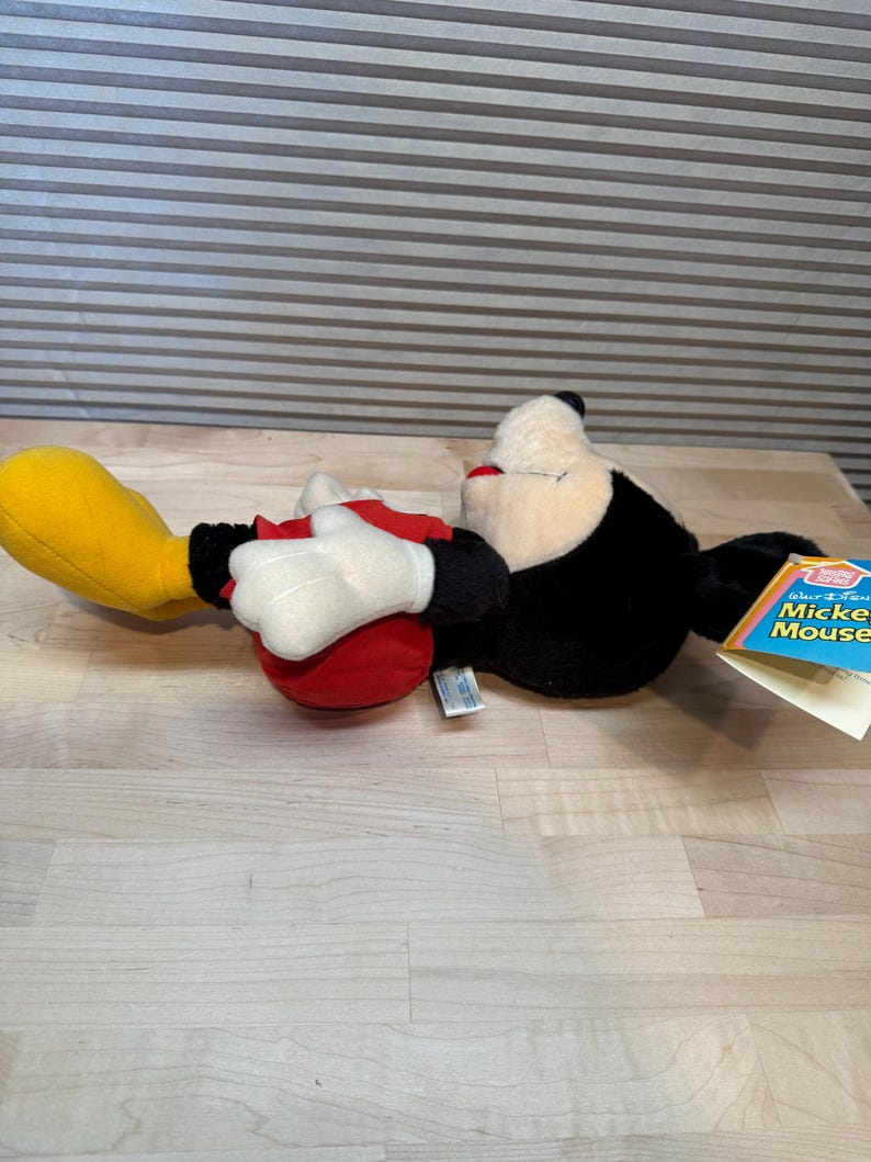 May include: A Mickey Mouse soft toy, lying on its side. The toy has a black body, white gloves, red shorts, and yellow shoes. A tag with the words "Walt Disney Mickey Mouse" is attached.