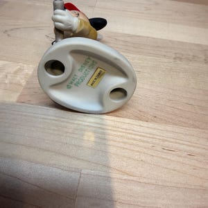 May include: A ceramic figurine of a Disney character, wearing a red hat and a yellow jacket, holding a pole. The base is off-white with the text "WALT DISNEY PRODUCTIONS" and "MADE IN TAIWAN".