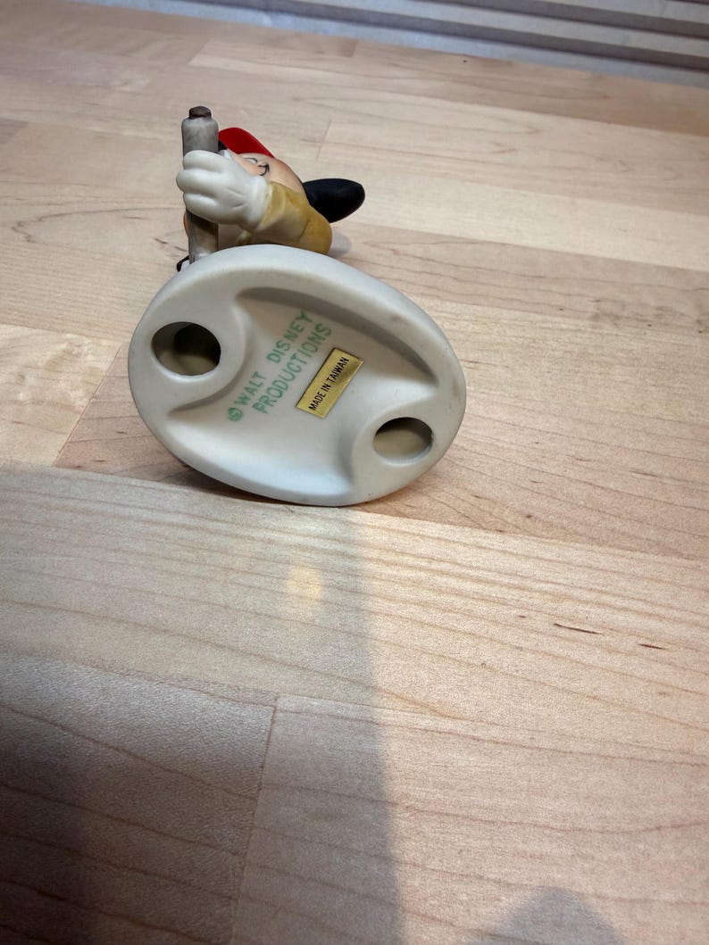 May include: A ceramic figurine of a Disney character, wearing a red hat and a yellow jacket, holding a pole. The base is off-white with the text "WALT DISNEY PRODUCTIONS" and "MADE IN TAIWAN".